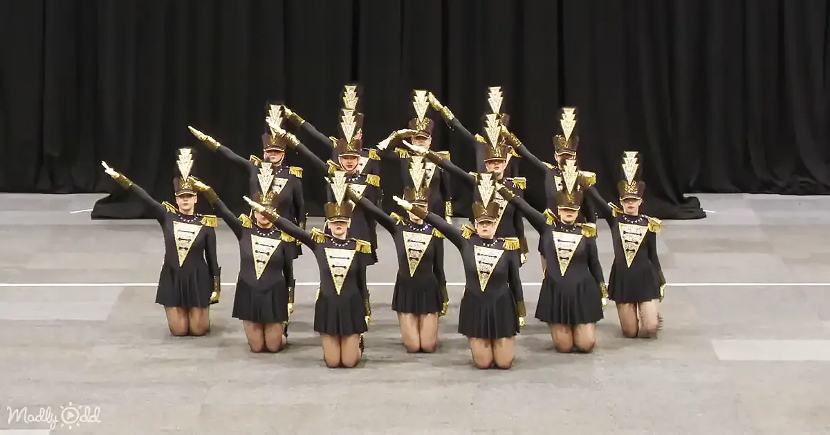 A synchronized dance team in black uniforms and gold accents performs a kneeling pose with arms raised.