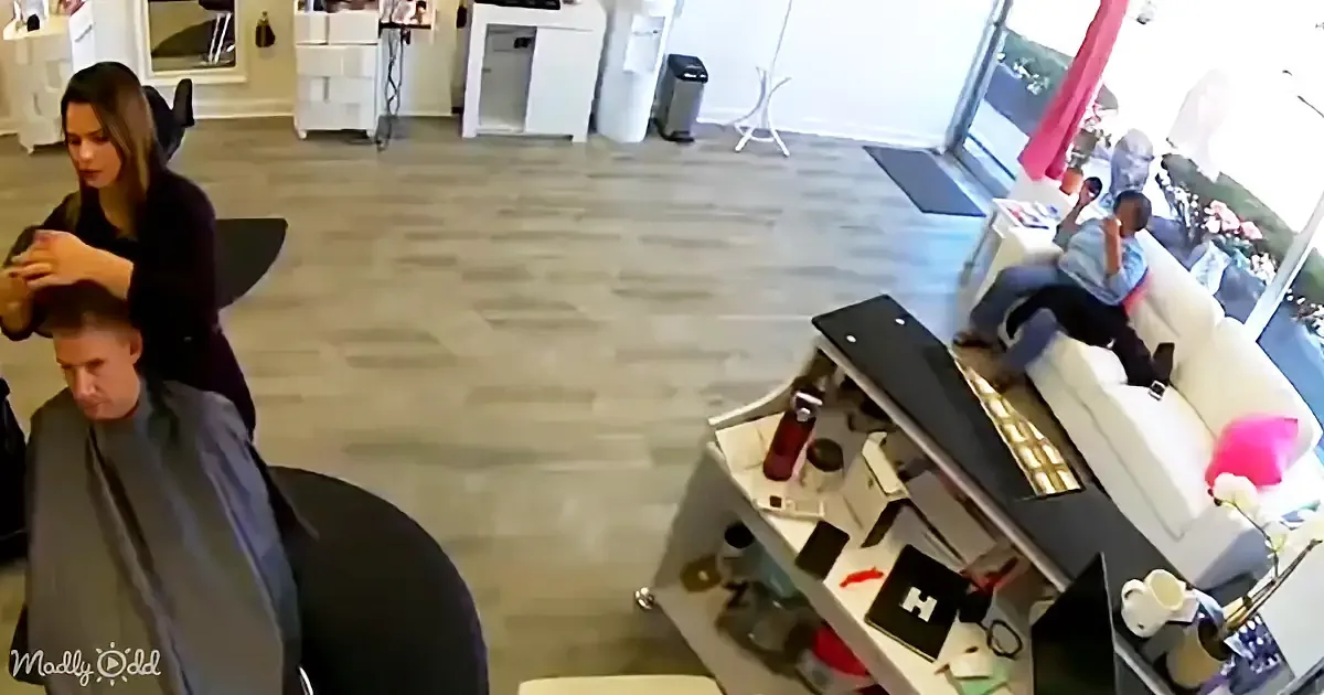 A woman is cutting a man's hair in a salon, with another person sitting on a couch in the background.