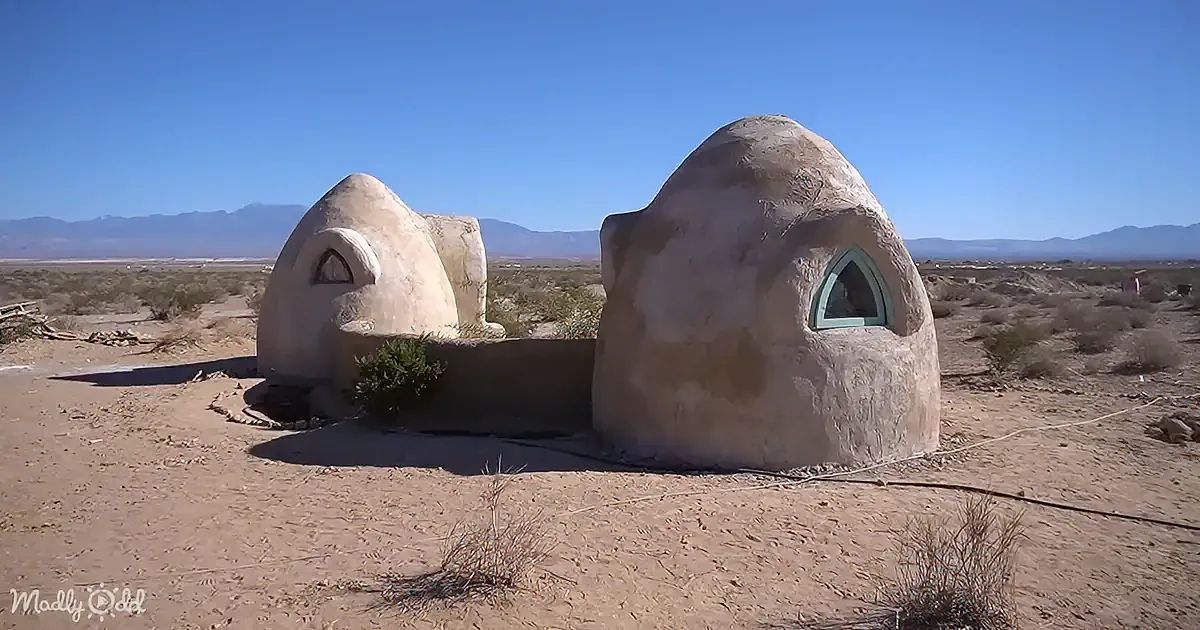 Two dome-shaped structures with arched windows stand in a desert landscape under a clear blue sky.