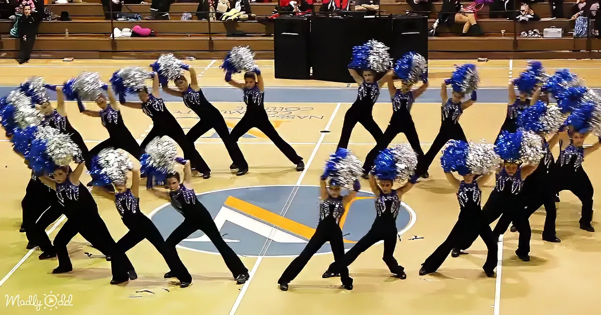 A synchronized cheerleading team in black uniforms and blue pom-poms performs a routine on a basketball court.