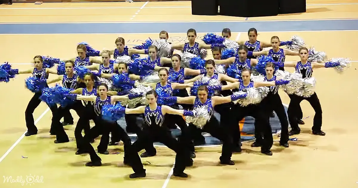 A synchronized cheerleading team in blue and black uniforms performs a routine on a gym floor.