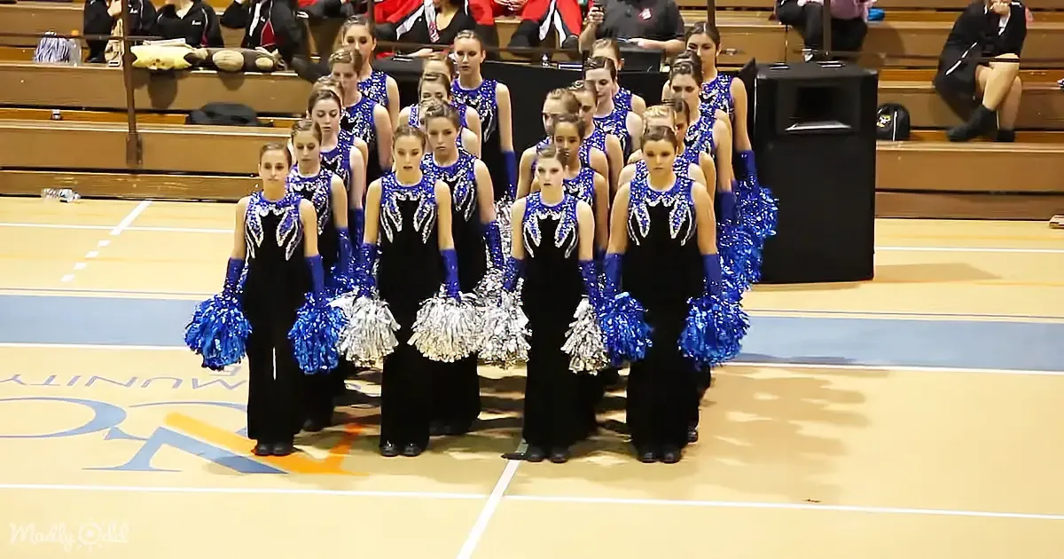 A synchronized dance team in blue and black costumes performs on a gymnasium floor.