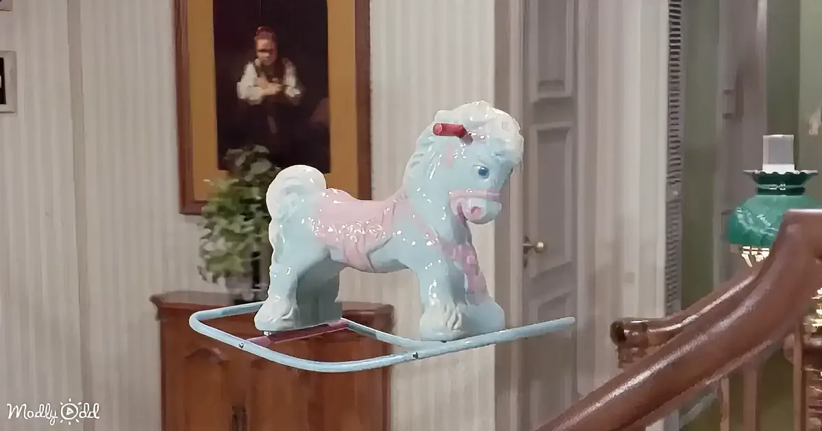 A light blue and pink rocking horse sits on a wooden stand in a room with a portrait and a green lamp.