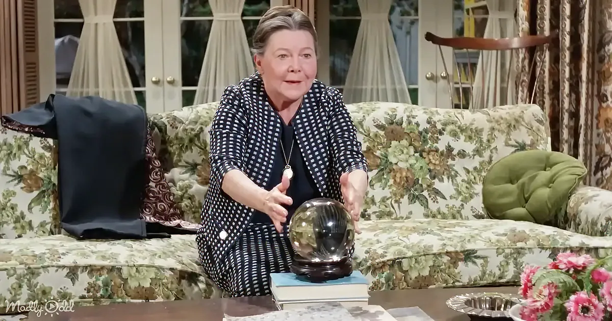 A woman in a polka-dot outfit sits on a floral-patterned couch, holding a crystal ball on a stand.