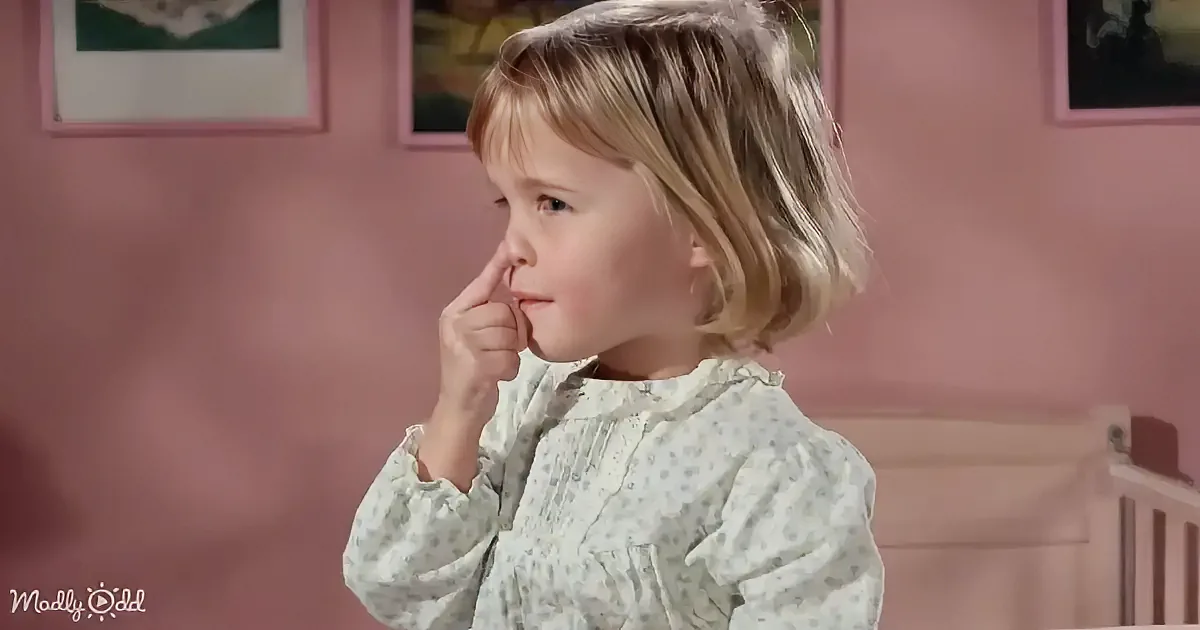 A young child with blonde hair, wearing a polka-dot shirt, is touching their nose in a room with pink walls and framed...