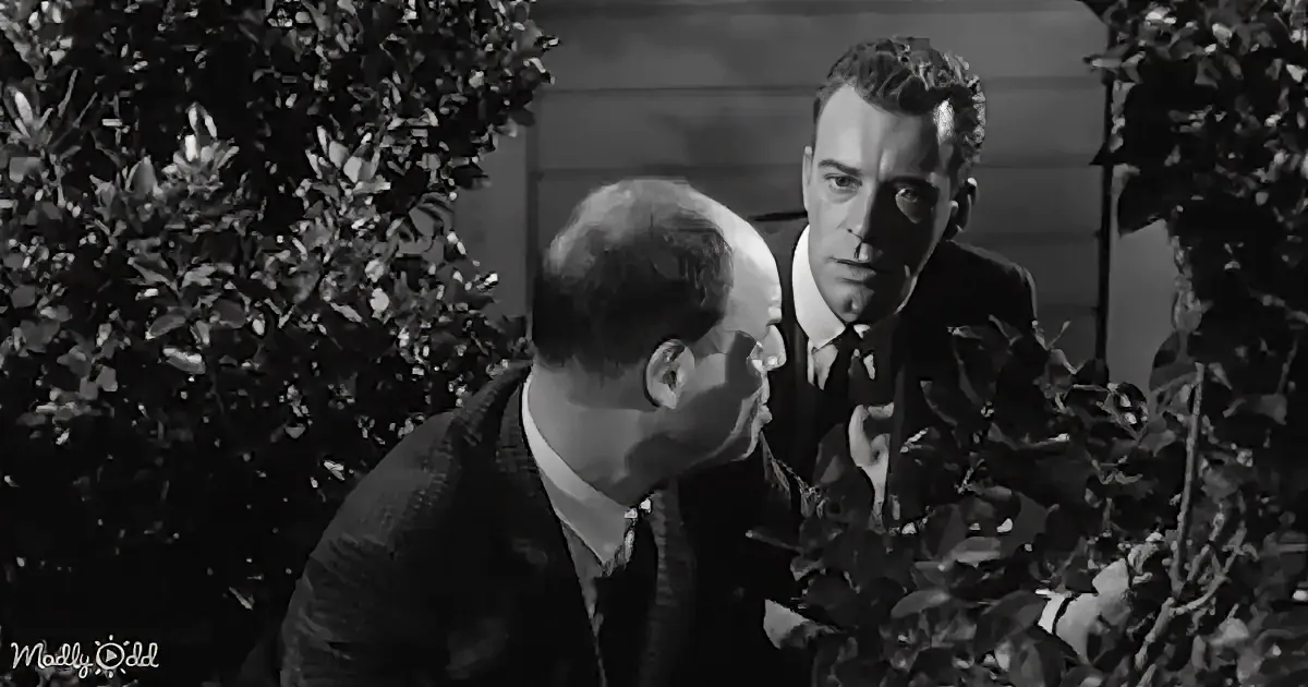 A man in a suit leans in closely to another man in a sweater, surrounded by bushes.