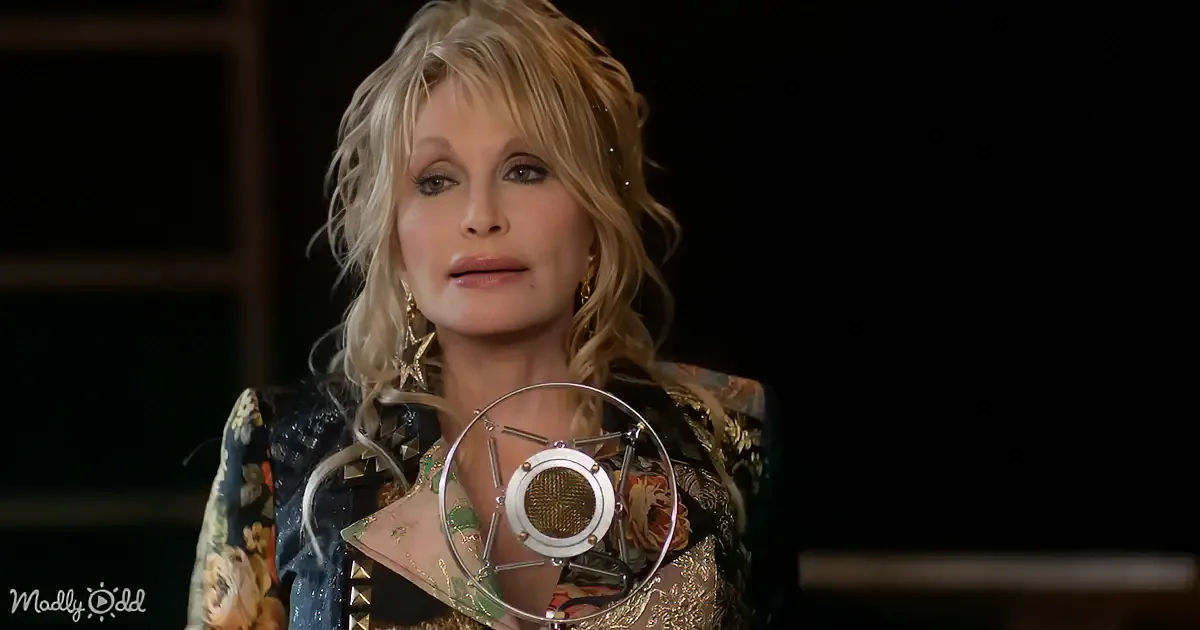 Dolly Parton is speaking into a microphone, wearing a floral jacket and star-shaped earrings.