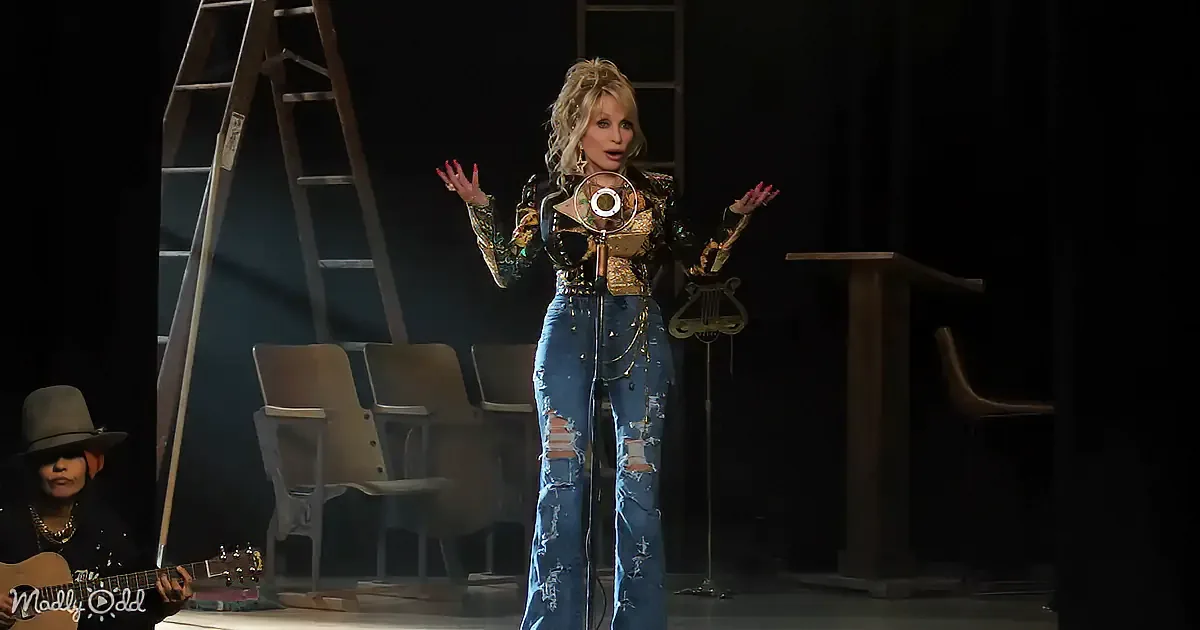 A performer in a glittery top and ripped jeans stands on stage, gesturing with hands near a microphone.