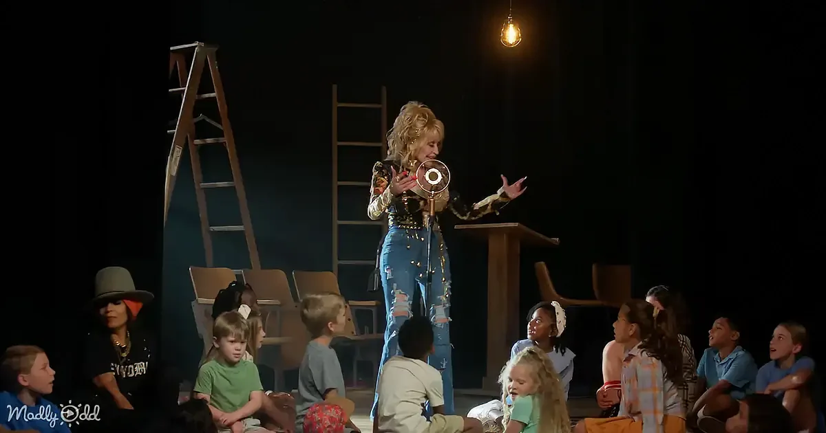A woman in jeans and a blouse stands on stage, holding a microphone, surrounded by children sitting on the floor.