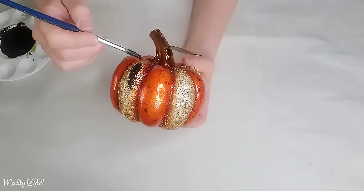 A hand holds a glittery pumpkin while painting it with a fine brush.