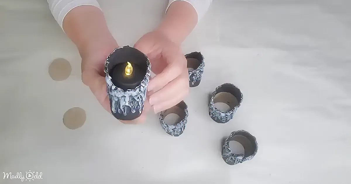 A hand holds a glowing candle with a black and gray textured holder, surrounded by similar holders on a white surface.