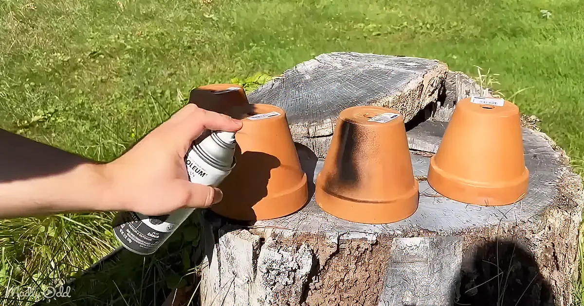 A hand holds a spray can, applying a substance to upside-down terracotta pots on a tree stump in a grassy area.