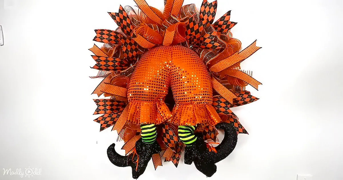 An orange Halloween wreath with a pumpkin face, black and orange checkered ribbon, and witch's boots.