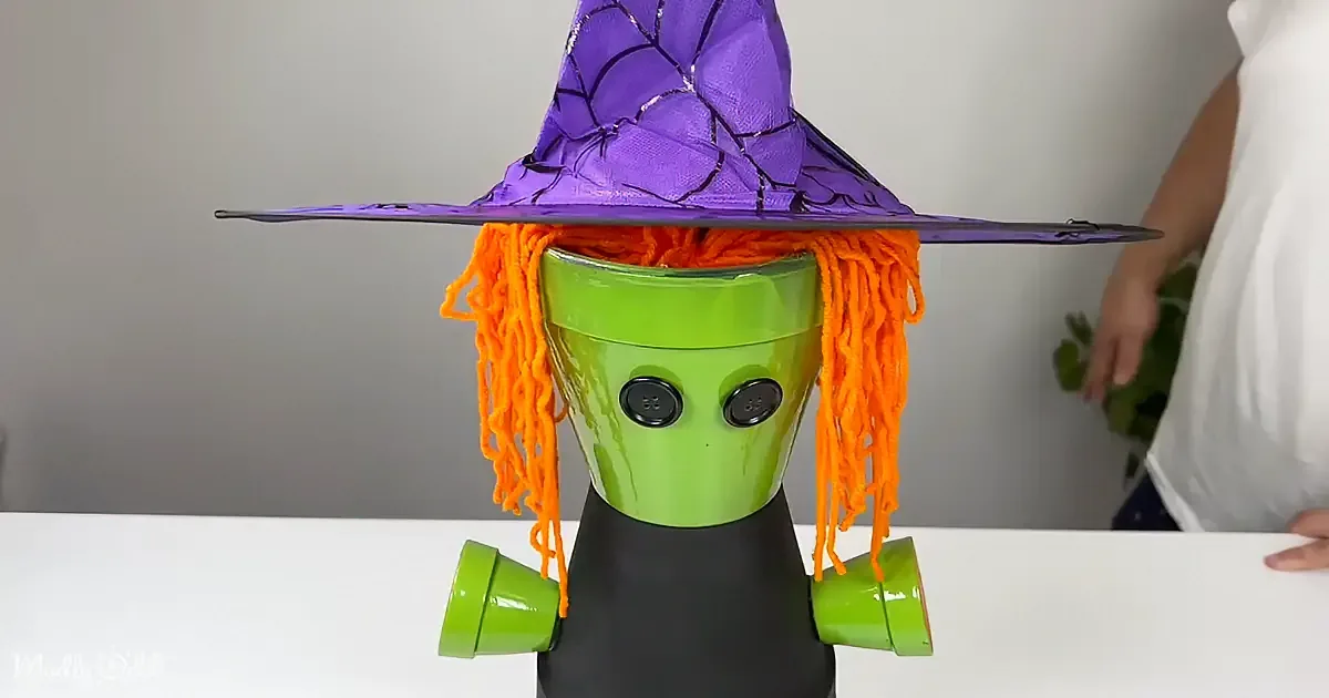 A whimsical witch figure made from plastic containers, adorned with a purple hat and orange yarn hair, sits on a white...