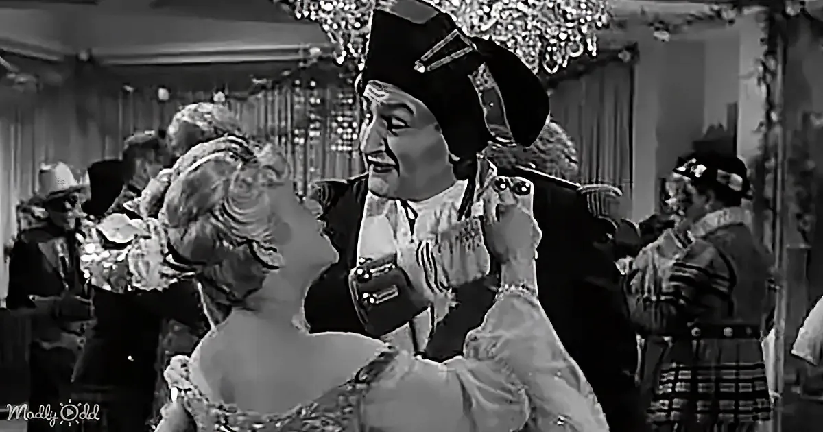 A man in a pirate costume dances with a woman in a formal dress, surrounded by other costumed figures in a grand hall.