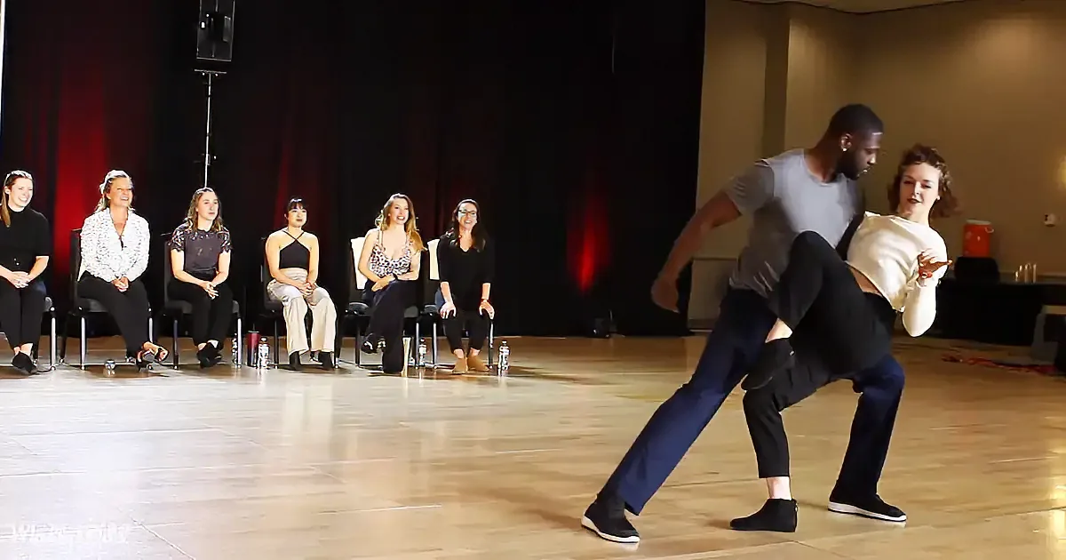 A man and woman dance on stage, with a row of seated women watching in the background.