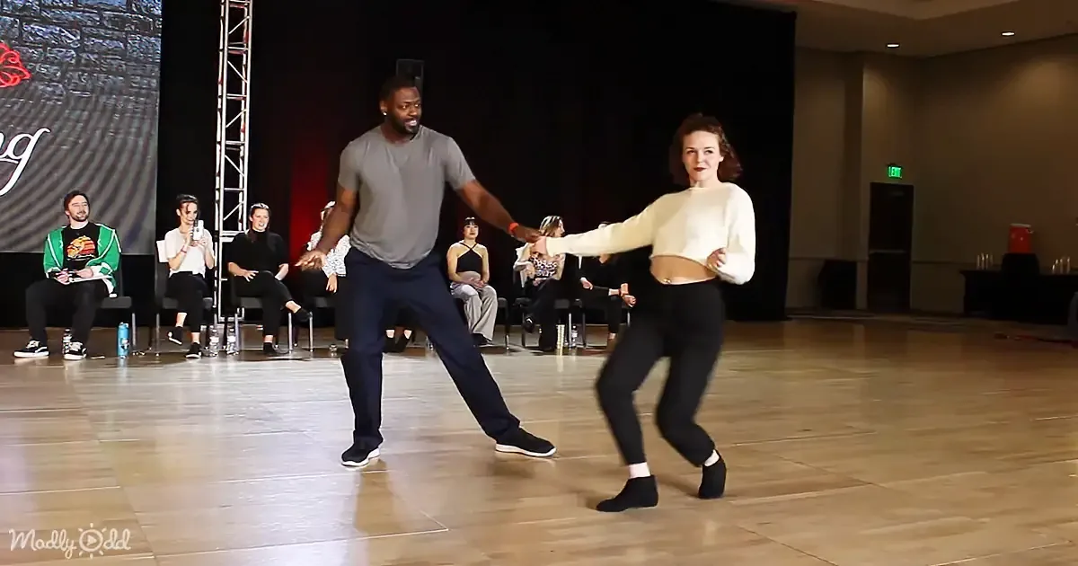 A man and woman dance together on a stage, surrounded by seated spectators.