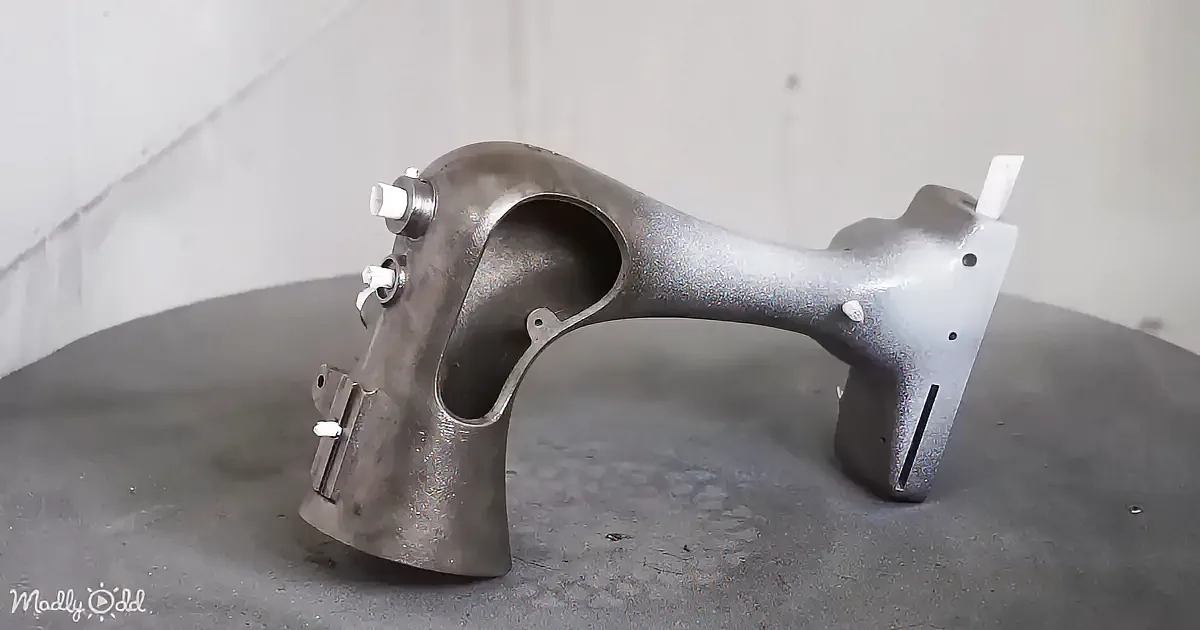 A metallic mechanical part with a curved design and multiple holes is displayed on a gray surface.
