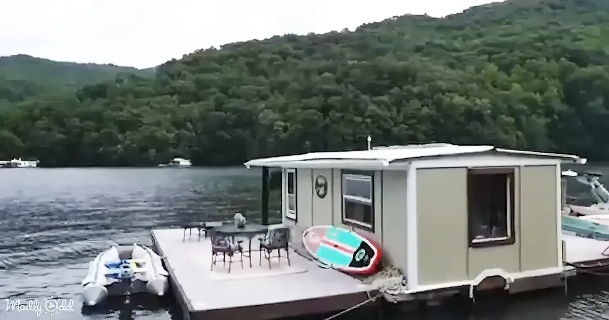A houseboat with a dining area, paddleboard, and motorboat docked on a lake surrounded by lush green hills.