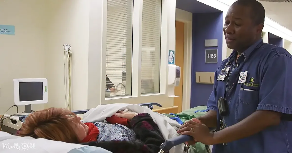 A man in blue scrubs stands beside a hospital bed, interacting with a patient.