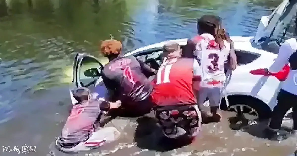 A group of people in football jerseys are gathered around a submerged car in a flooded area.