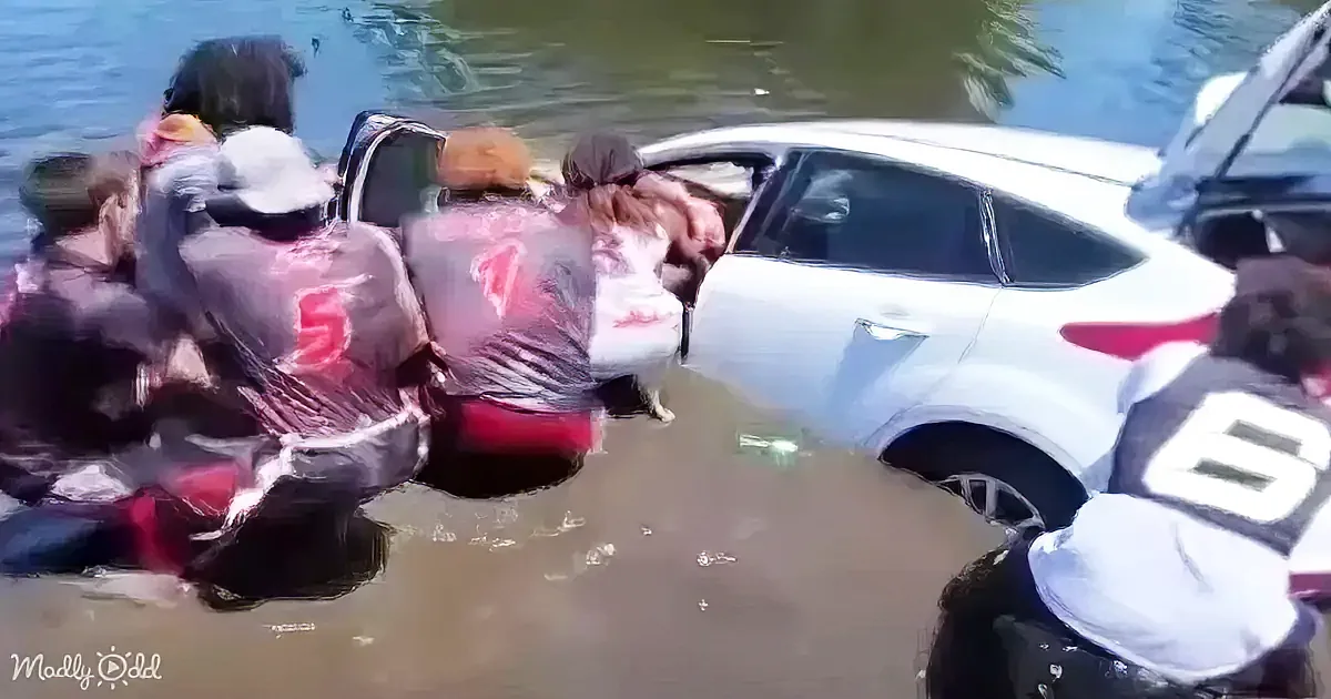 A group of people, some in life jackets, are assisting a white car stuck in floodwaters.