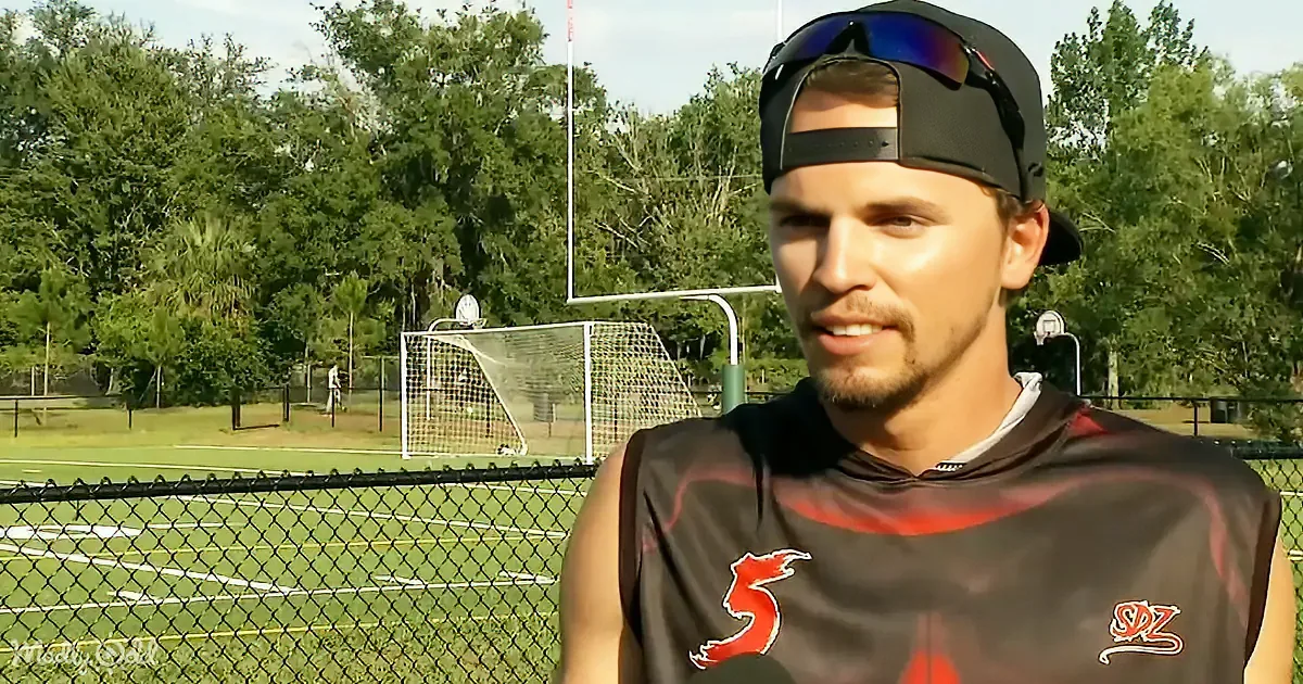 A man in a black jersey with a red number 5 stands in front of a chain-link fence and a soccer field with trees in the...