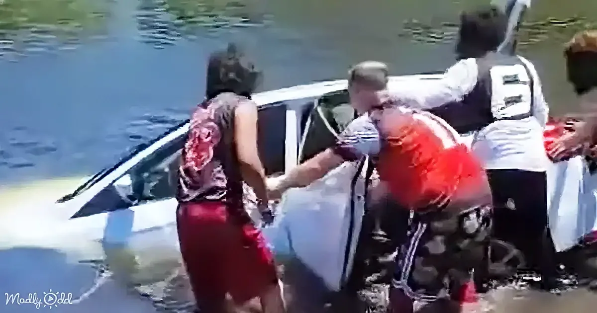 A person in a red dress is helping another person out of a submerged car in a body of water.