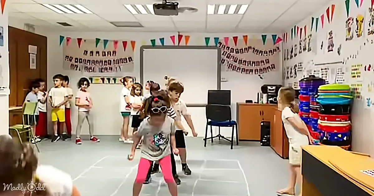 A girl in a classroom, wearing a hat and goggles, is dancing in front of a group of children.