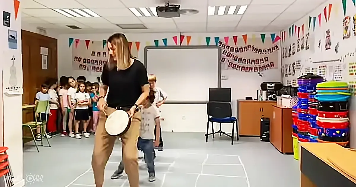 A woman plays a drum in a classroom while children watch, with colorful decorations and a whiteboard in the background.