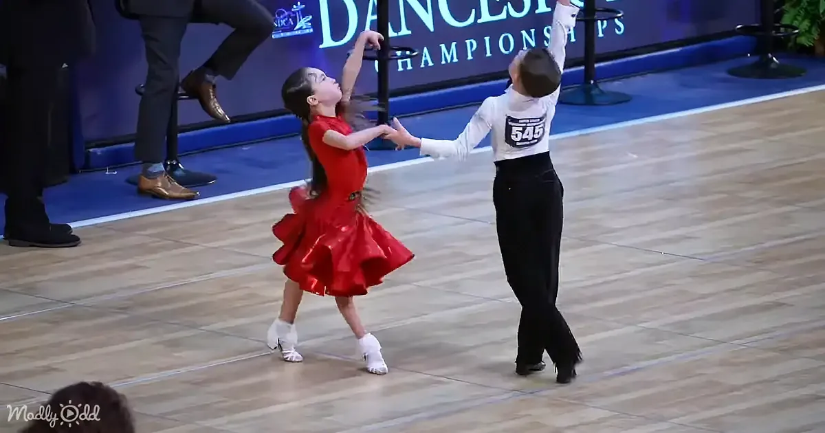 A young girl in a red dress dances gracefully with a partner in a ballroom competition.
