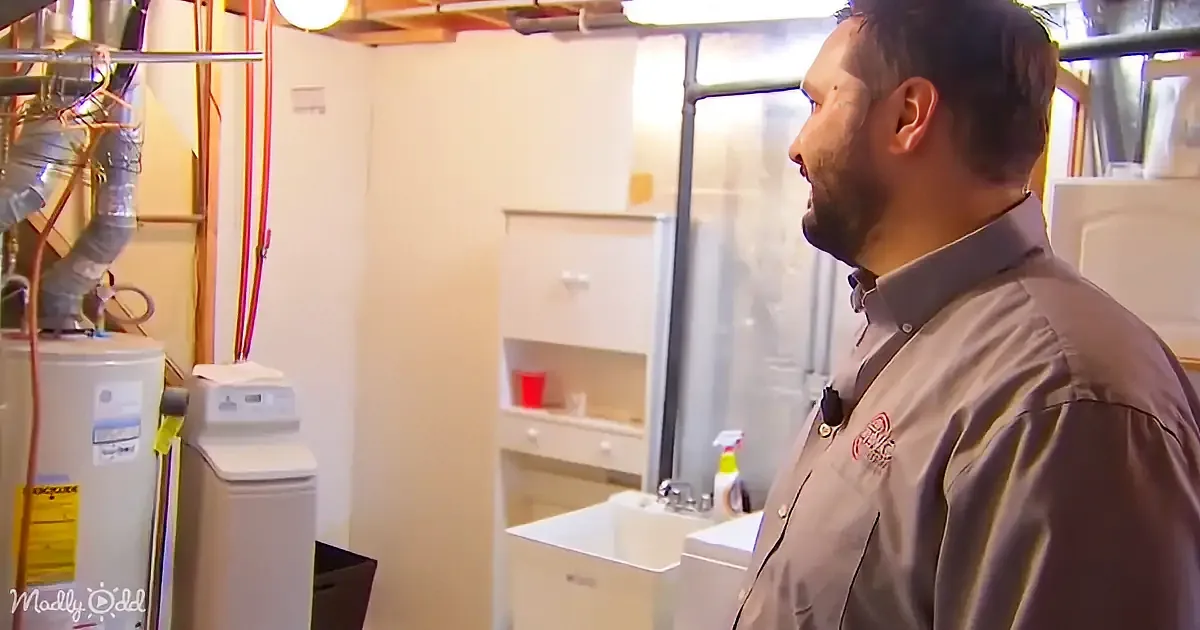 A man in a uniform stands in a utility room with visible pipes, a water heater, and a sink.