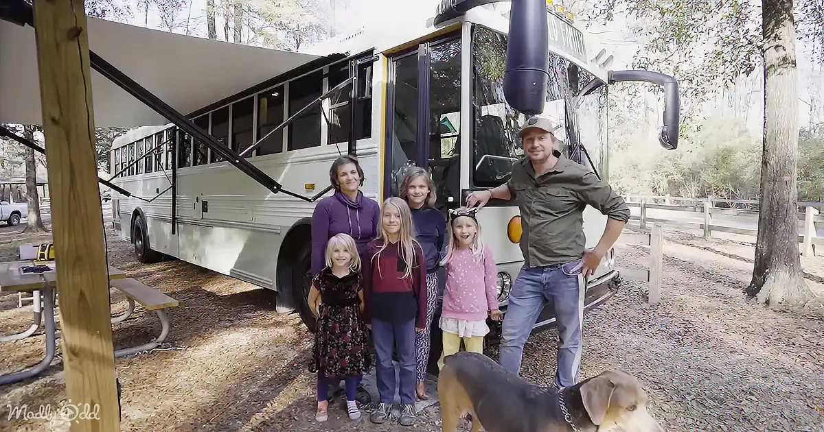 A family stands in front of a converted bus, surrounded by trees and a picnic table, with a dog nearby.