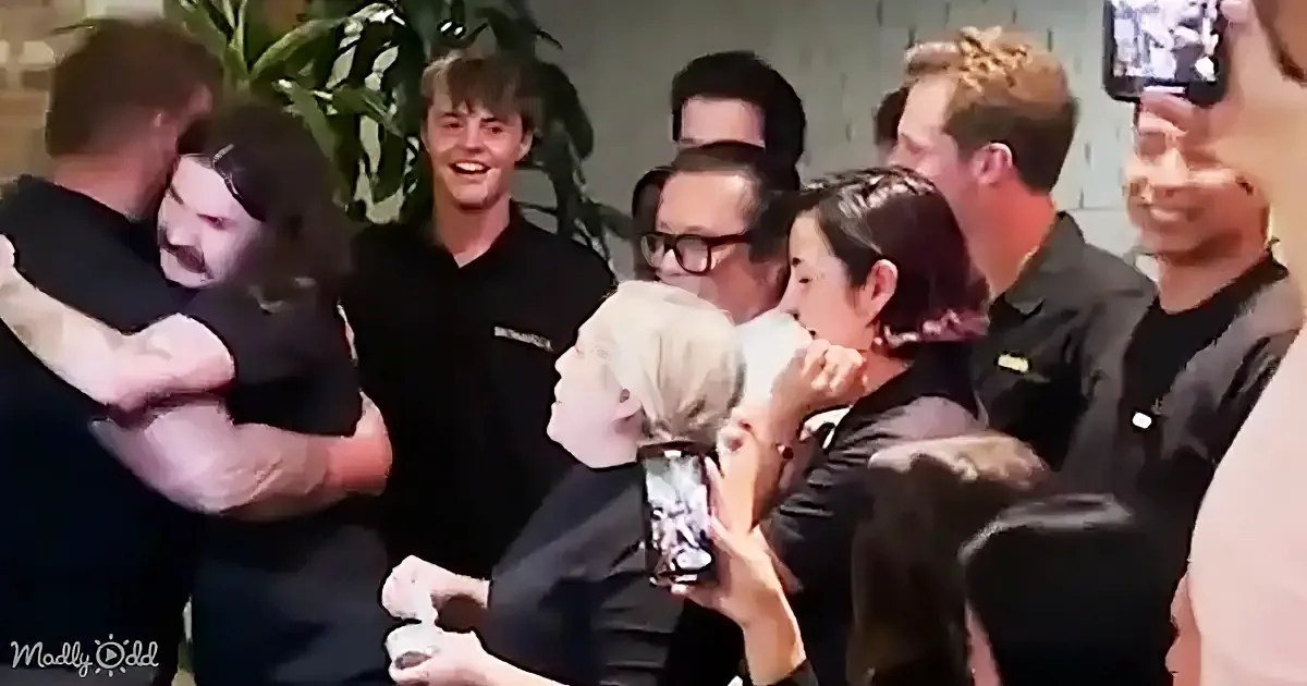 A group of people, mostly in black, are gathered, with one person hugging another, while others watch and smile.