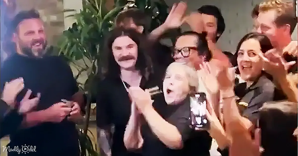 A woman in a black shirt claps enthusiastically, surrounded by a group of people also clapping and smiling.