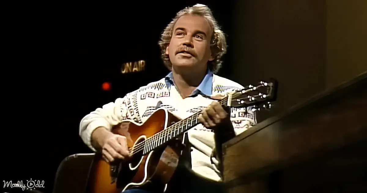 A man with a mustache plays an acoustic guitar on a stage with an "On Air" sign in the background.