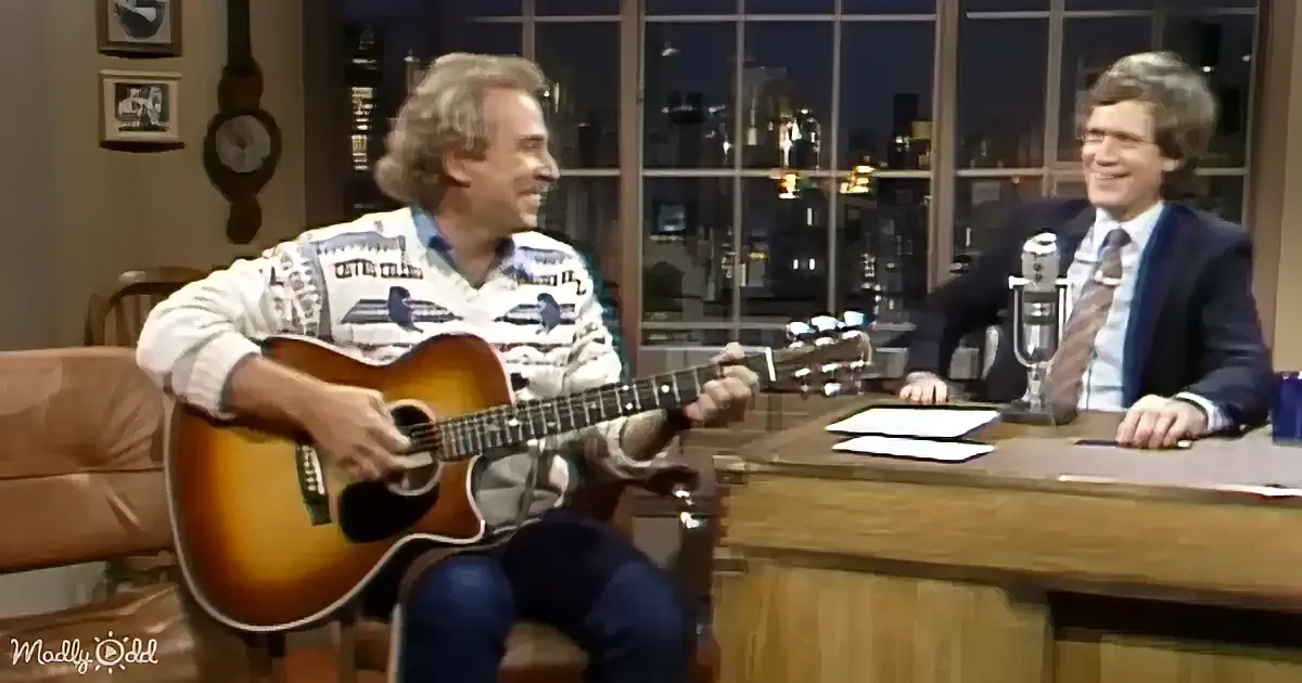 A man in a patterned shirt plays guitar while another man in a suit sits at a desk, both smiling.
