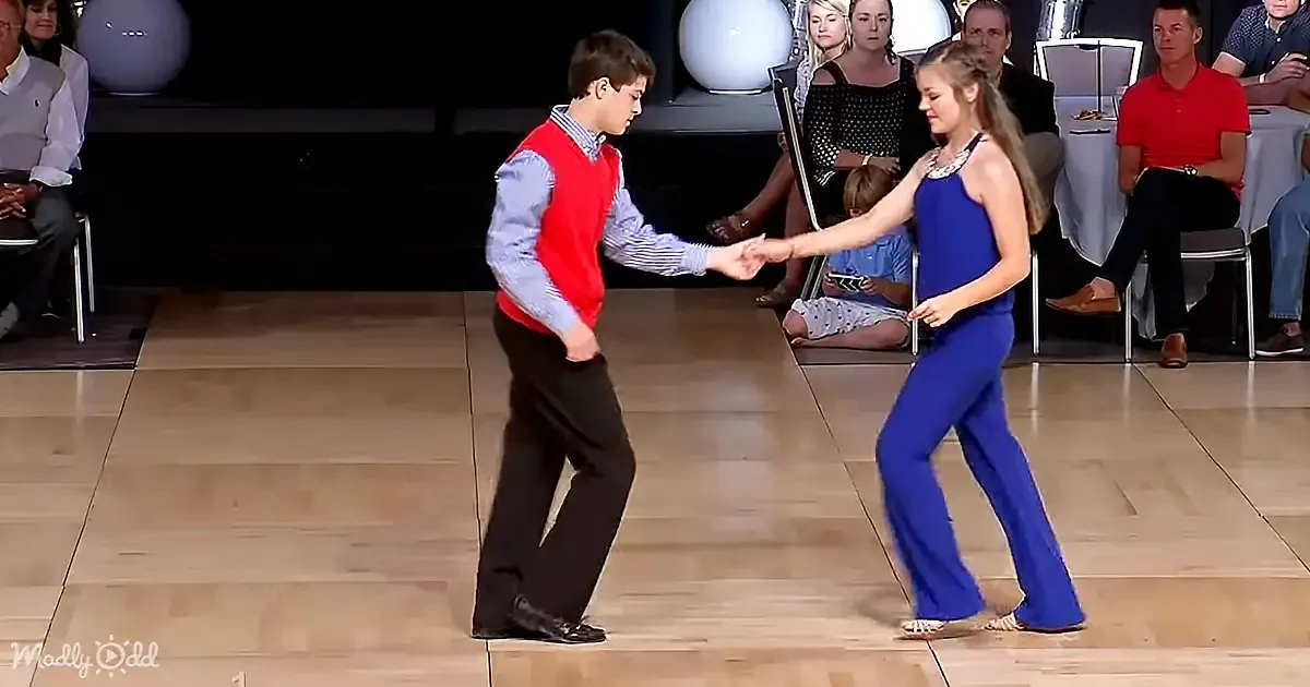 A boy and girl in formal attire dance on a wooden floor, surrounded by spectators.