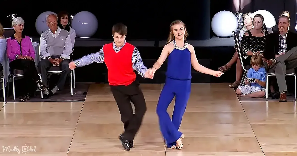 A boy in a red vest and a girl in a blue dress dance together on a wooden floor, surrounded by seated spectators.