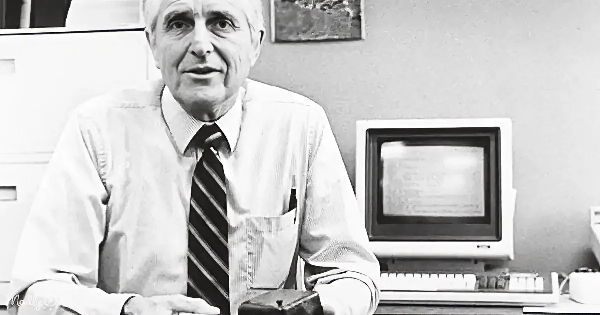 An older man in a shirt and tie sits at a desk with a computer, appearing to speak.