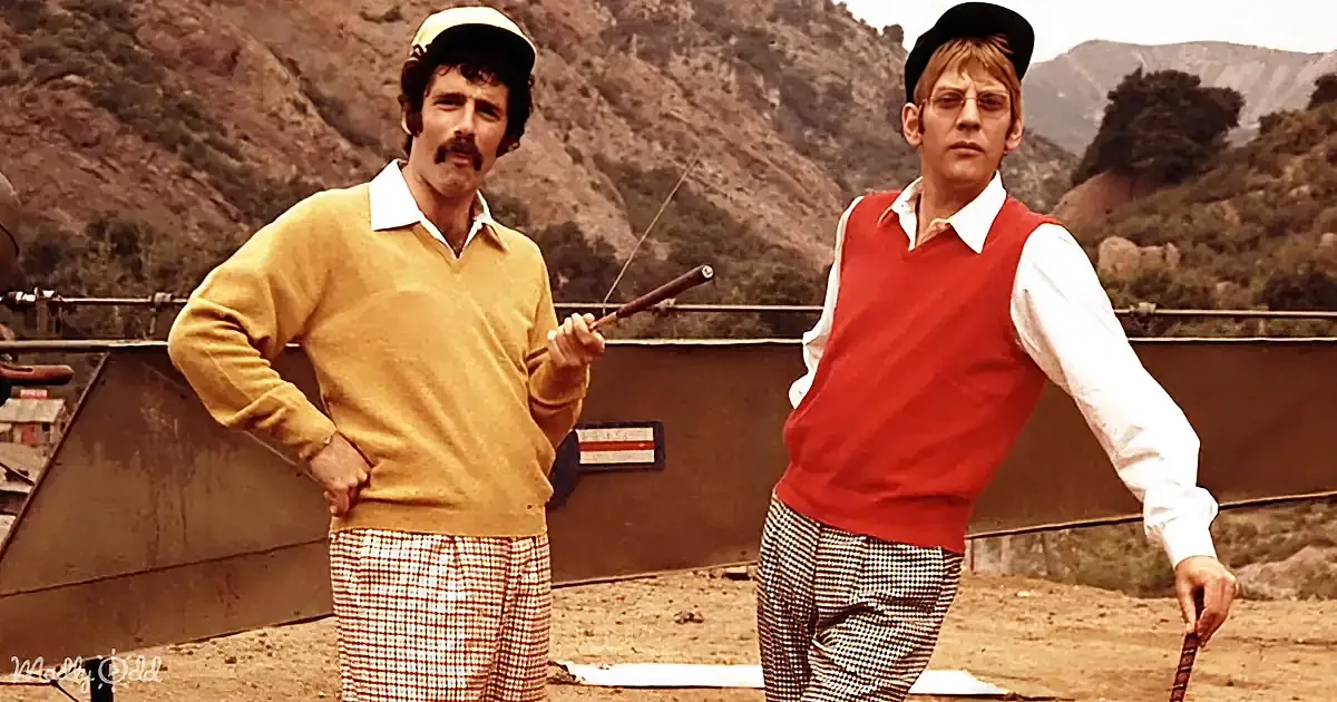 Two men in vintage clothing stand outdoors, one holding a stick, against a mountain backdrop.
