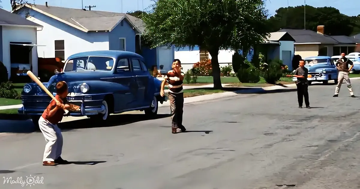 A boy swings a bat in a street scene with vintage cars and houses in the background.