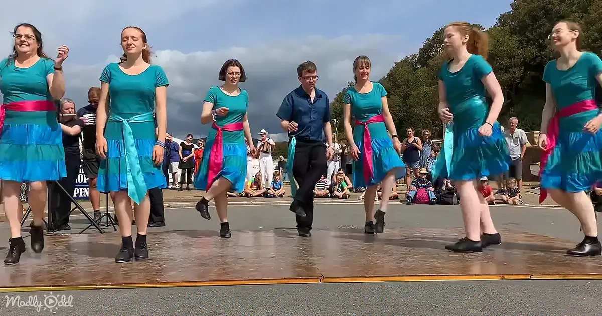 A group of dancers in teal dresses with pink sashes performs on a stage in front of an audience under a partly cloudy sky.