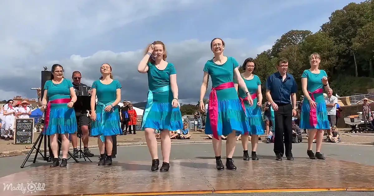 A group of dancers in teal tops and blue skirts perform on a stage under a cloudy sky.