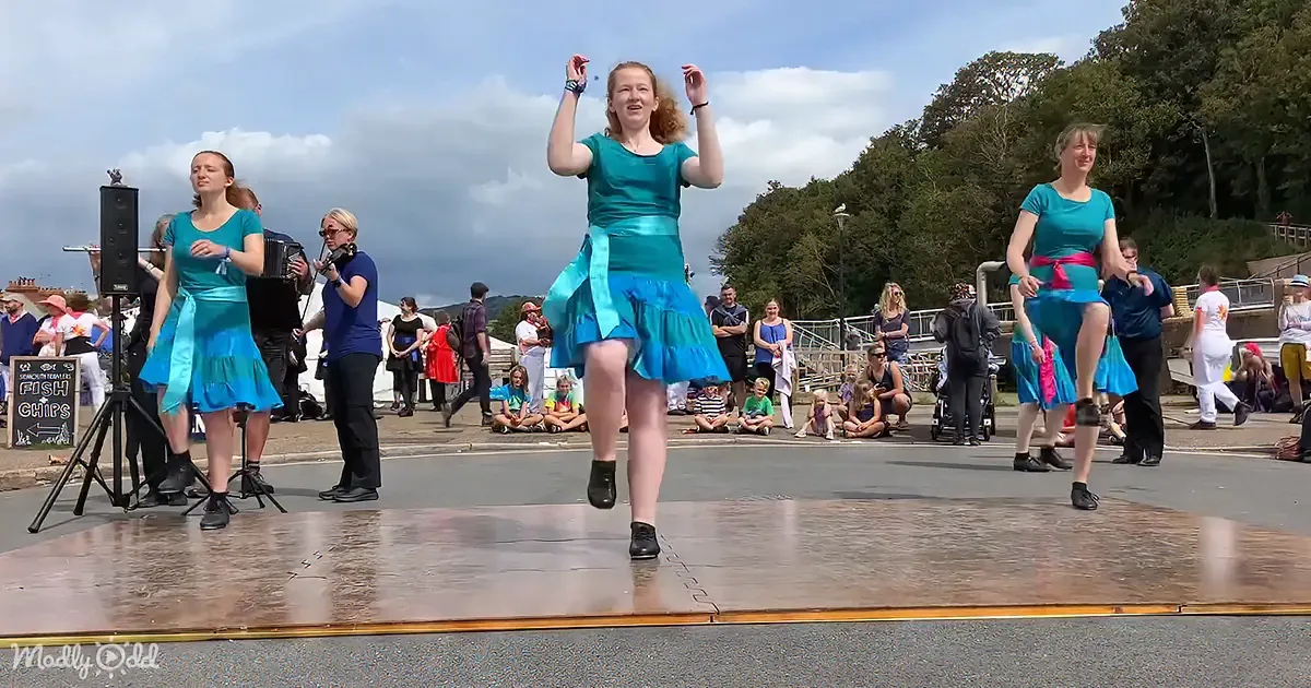 A woman in a teal dress dances on a stage with others, surrounded by an audience and a cloudy sky.