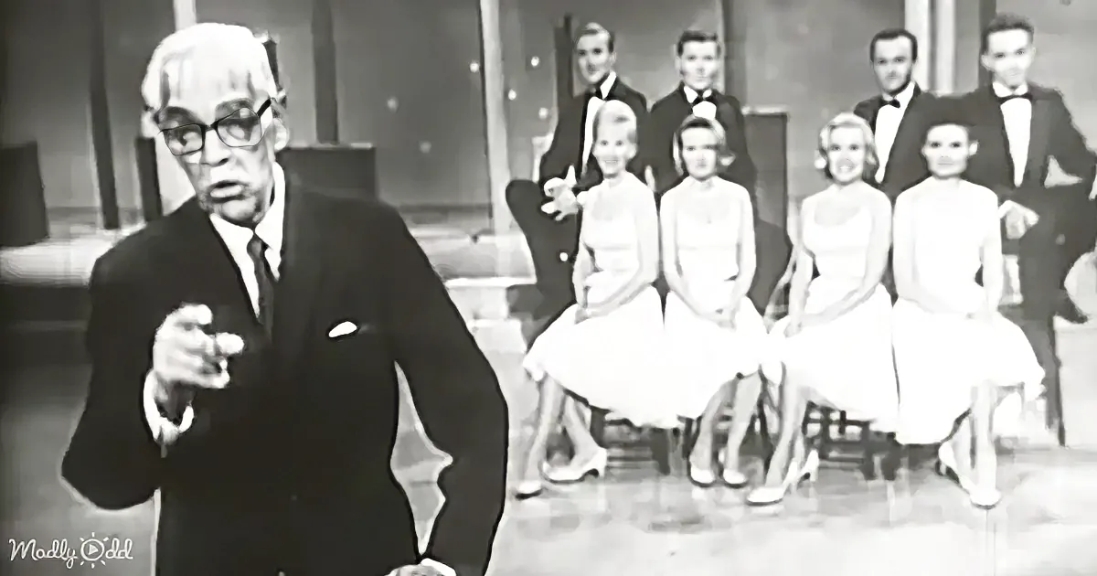 A man in a suit gestures while seated women in white dresses and men in tuxedos watch behind him.
