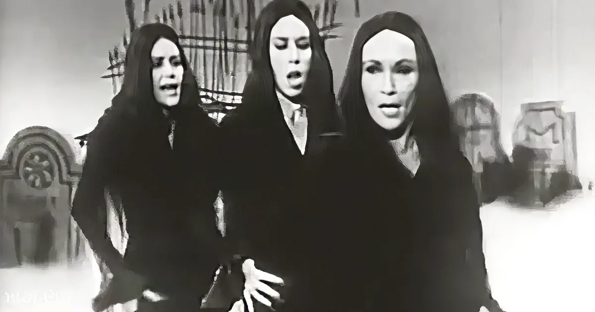 Three women in black robes stand together, mouths open, in a monochromatic setting.