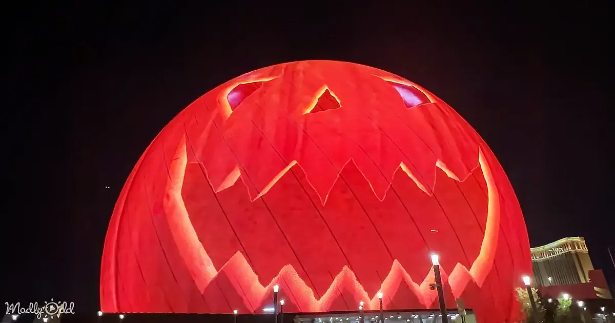 A giant illuminated red pumpkin with a carved face stands prominently against a dark sky.
