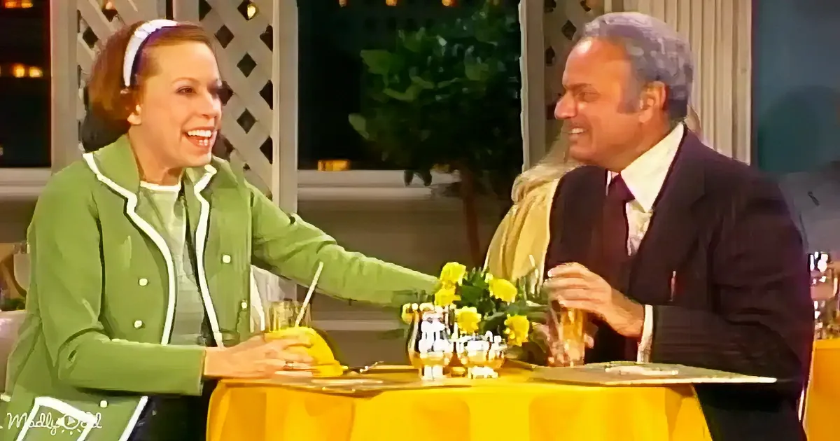 A woman in a green jacket and a man in a suit are seated at a table with yellow flowers, smiling and interacting.