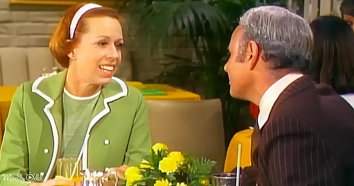 A woman in a green outfit smiles at a man in a pinstripe suit at a table with yellow flowers.